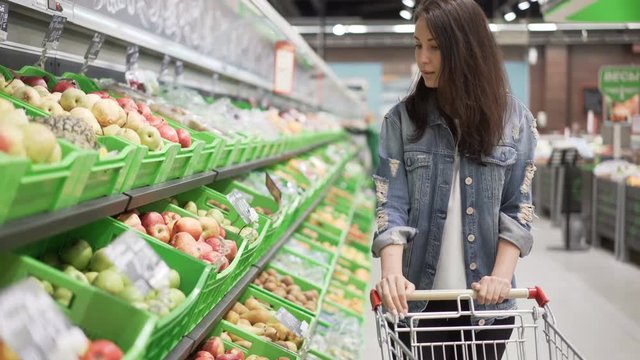 Pretty young woman is choosing fruit in grocery store, she is touching and smelling apples then putting them in trolley. Healthy food, beautiful girls and shops concept.