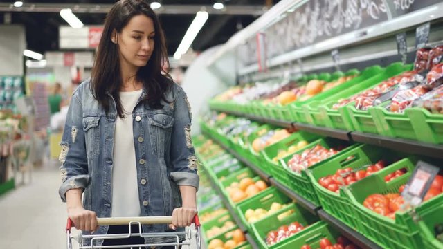 Pretty young woman is walking along fruit and vegetable row pushing shopping trolley and looking at organic food with smile. Healthy lifestyle and supermarket concept.