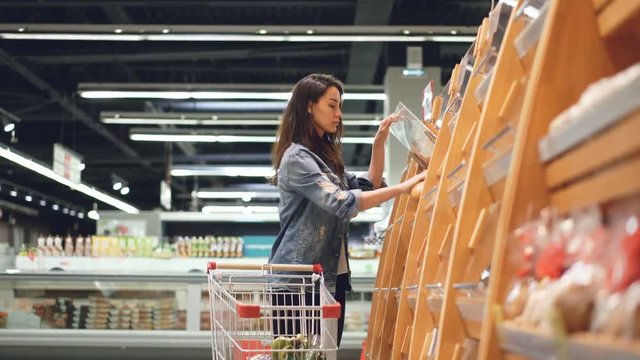 Attractive girl customer is buying bread in bakery department is shop, smelling it, smiling and putting in shopping trolley. Healthy lifestyle and supermarket concept.