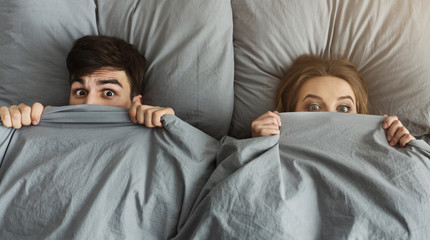 Shocked man and woman hiding under blanket in their bed at home