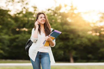 A young girl student with notebooks and a folder with headphones in her ears listening to an online...