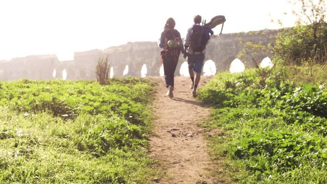 Young lovely couple backpackers tourists walking holding hands toward roman aqueduct arches in parco degli acquedotti park ruins in rome on romantic misty sunrise with guitar and sleeping bag slow