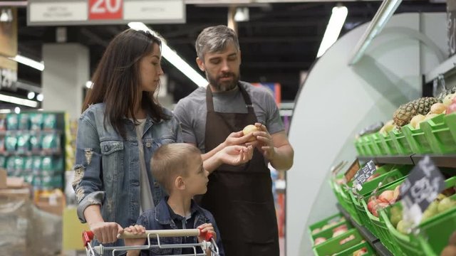 Experienced shop assistant is selling fruit to cheerful young family mom and child, giving them pears, apples and pineapple, happy customer is laughing and smiling.