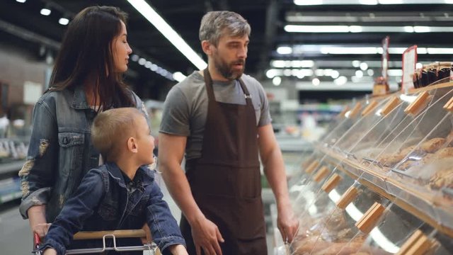 Shop assistant is working in bakery department in supermarket then packing bread roll for nice family mother and son and giving food to attractive woman.
