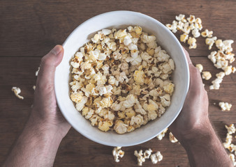 top view of hands holding bowl filled with popcorn above wooden table