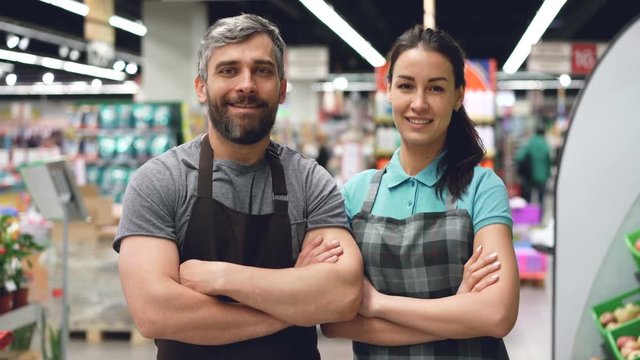 Portrait of two supermarket employees attractive people in aprons standing inside shop, smiling and looking at camera. Shelves with food and drinks are visible.