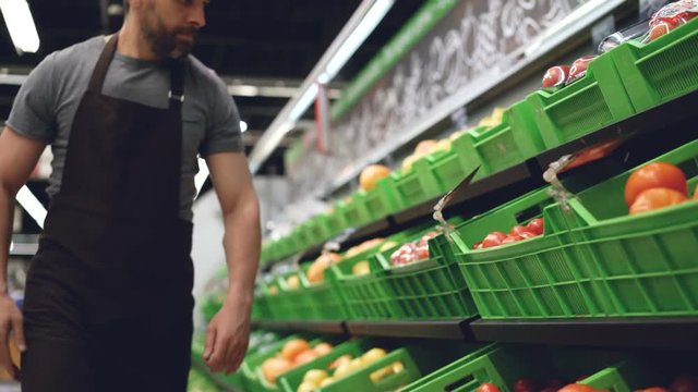 Strong salesman wearing uniform is bringing plastic box of tomatoes and putting on shelf in supermarket in fruit and vegetables department. Containers with food are visible.