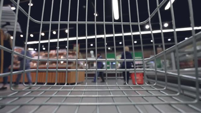 Shopping trolley is moving quickly along aisles in supermarket, shelves with food, drinks and goods are visible through cart grille. Shopping and customers concept.