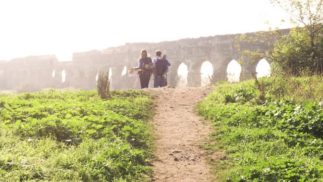 Young lovely couple backpackers tourists walking holding hands toward roman aqueduct arches in parco degli acquedotti park ruins in rome on romantic misty sunrise with guitar and sleeping bag slow