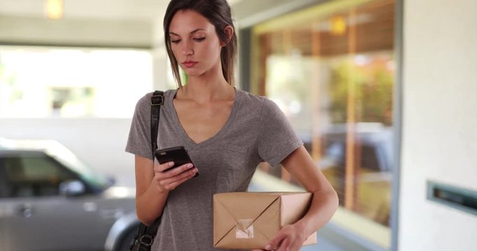 Woman using mobile phone while holding mail package standing outside her house, Millennial female using smartphone app to track package before leaving for post office, 4k