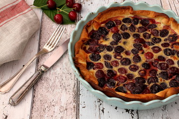 Fresh homemade cherry pie in blue baking pan with some raw cherries and fork and knife on linen kitchen towel, wooden background