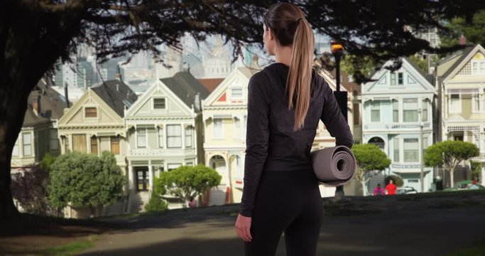 Fit young woman carrying yoga mat walking onscreen near painted ladies houses in San Francisco, Rear view of yoga girl entering shot and walking around in San Francisco park, 4k