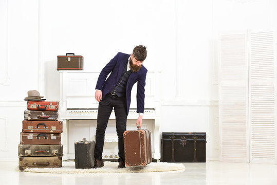 Macho elegant on strict face stands near pile of vintage suitcase, ready for vacation. Man, traveller with beard and mustache with luggage, luxury white interior background. Luggage and travel concept