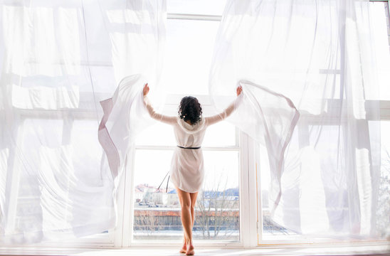 the young woman stands on the window among the open curtains