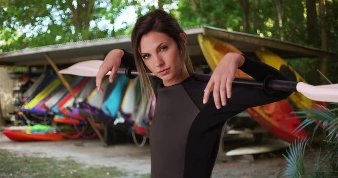 Young woman in wetsuit holding kayak paddle over shoulders near kayaks in outdoor setting, Sporty female athlete in her 20s with wetsuit and oar posing by shack of kayaks, 4k