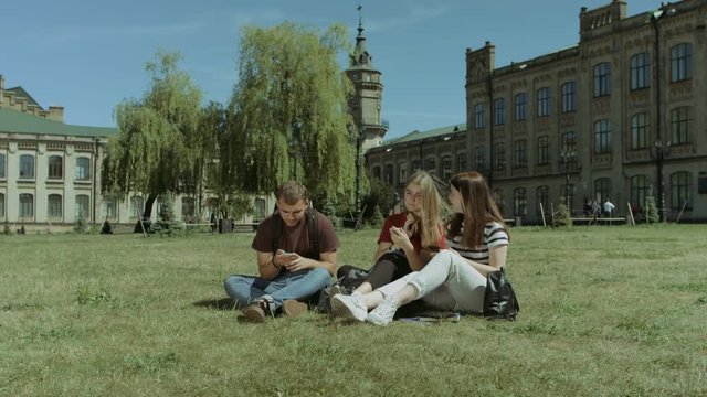 Group of smiling college students browsing social networks on smart phones and chatting while sitting on university campus lawn. Positive students networking with cellphones and talking on park lawn.