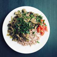 Red rice with mushrooms and cherry tomatoes. Vegan dish. European cuisine. Simple healthy lunch on the black background. Vegetarian rice dish on the white plate. Close up. Toned photo. Top view.