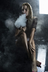 Fototapeta na wymiar Beautiful topless athletic big breasted blonde girl wearing white sexy bodysuit and mesh transparent top vaping sitting on wet floor in scenic smoke under falling water drops on black.