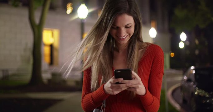Millennial woman texting on smartphone looking happy outside in the evening, Caucasian girl in her 20s happily texting on phone standing on sidewalk outside, 4k