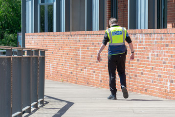 security guard in yellow hi vis with walkie taklie walking away from job on wooden decking by brick wall after being fired with head bowed