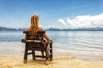 Fototapeta premium A girl sitting on a bamboo chair on the beach on a blue sea and sky background. place for text.