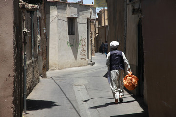 mężczyzna idący wąską boczną uliczką między domami w słoneczny dzień w iranie - obrazy, fototapety, plakaty