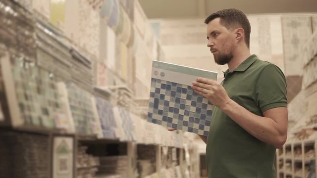 Shopper man is standing in trading area of huge retail shop with building materials. He is looking samples of ceramic tiles for walls
