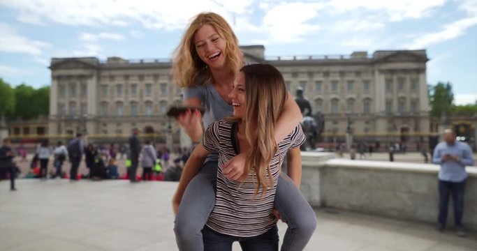 A pair of playful white girl friends playing piggyback and taking selfies together outside in London, Blonde female jumps on attractive brunette friend's back to take selfies in London, England, 4k