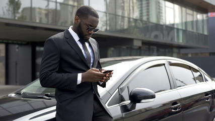 Fototapeta na wymiar Confident man typing e-mail on smartphone, standing near his luxury car