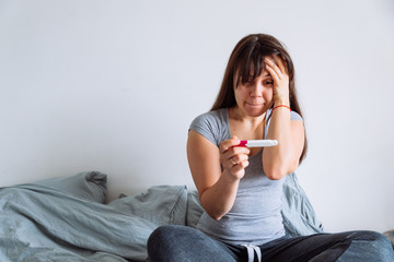 woman with worried view looking at pregnancy test while sitting in bed