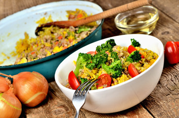 Close-up of fork and tuna risotto with vegetables, tomatoes, broccoli and parsley in the bowl, onions, oil and pan in the background