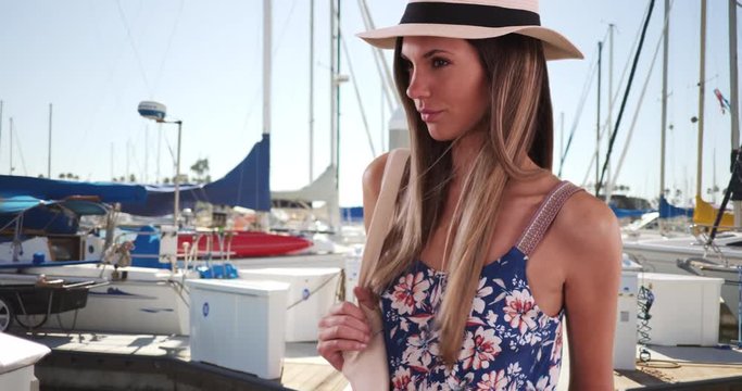 Pretty lady in floral romper wearing fedora and holding bag by some docked boats, Stylish young woman in her 20s in summer dress standing by boat dock outdoors, 4k