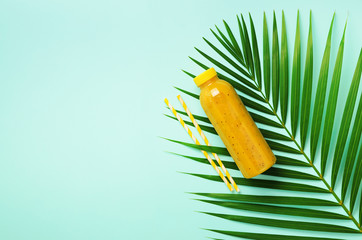 Palm leaves and bottle of orange, pineapple, mango smoothie, straw on blue background. Detox summer drink. Vegetarian concept. Top view, flat lay, copy space