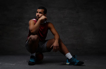 Naklejka premium Pensive young African-American basketball player in sportswear sitting on a ball over dark background.