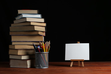 A stack of books and a stand for pens on a table, on a black background. A small easel, a white blank, a blank, a mock-up.
