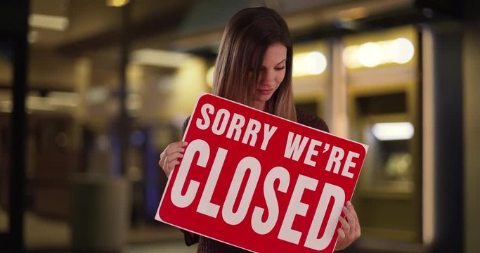 Caucasian Woman holding Closed sign standing by storefront at night, Female looking at camera with Sorry We Are Closed sign outside store at night, 4k