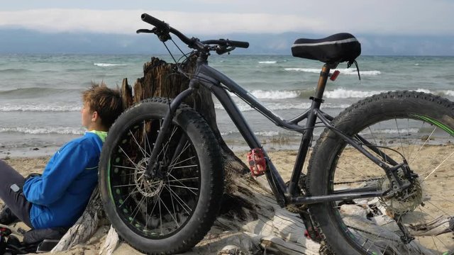 Fat bike also called fatbike or fat-tire bike in summer driving on the beach. The guy and his bike rest on the sandy beach. He sits and rests after a long trip. Enjoys the beautiful sea view and the