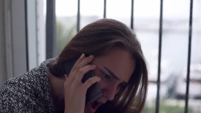 Close-up of depressed woman is talking over the mobile phone, yelling and crying sitting on the floor by the window with rain falling outside. Lonely woman in despair with the suicide thoughts.