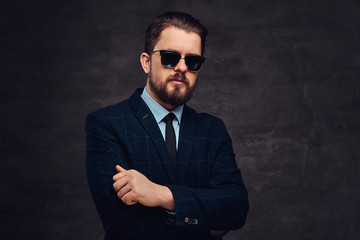 Close-up portrait of a handsome fashionable middle-aged man with beard and hairstyle dressed in an elegant formal suit and sunglasses. Isolated on a textured dark background in studio.