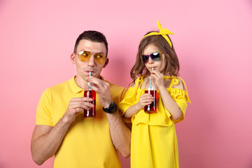 Crop view of young man and cute child in yellow outfits holding red drinks with straws looking at camera and smiling on pink background