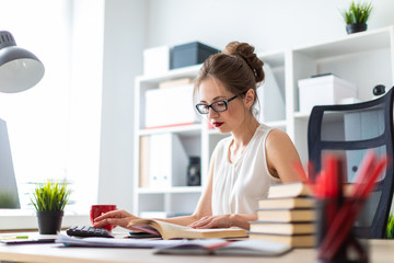 A young girl sits at a computer table and holds an open book in her hands and prints on the keyboard.