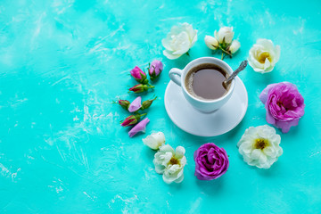 Tasty morning concept. Flat lay of cup of freshly brewed coffee surrounded with white and pink roses flowers and petals over light green pastel background, cozy breakfast.copy space