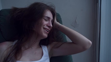 Slow motion of young depressed woman in trouble pulling her hair with hands and crying sitting in chair. Lonely woman in despair with suicide thoughts.