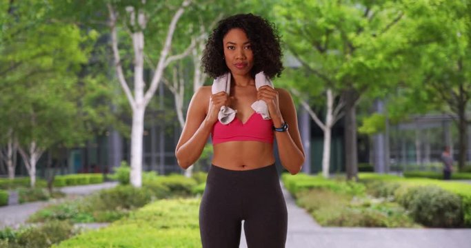 Smiling African American woman after workout standing in front of trees and large building, Runner or jogger after finishing work out in gym stands with towel around neck, 4k