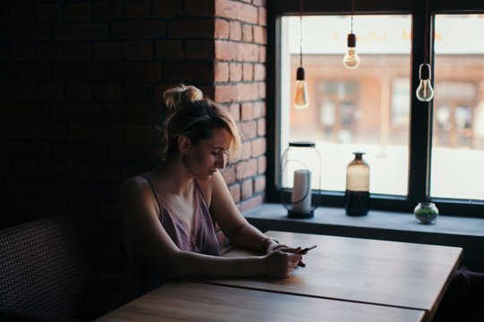 Side view portrait of young female using her smartphone sitting at the table at cafe indoors