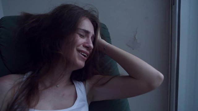 Slow motion of young depressed woman in trouble pulling her hair with hands and crying sitting in chair. Lonely woman in despair with suicide thoughts.