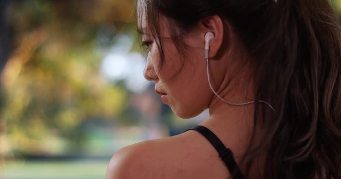 Tight shot of millennial female runner with confidence standing outdoors at park, Profile view of young Asian athlete wearing earbuds out in nature, 4k