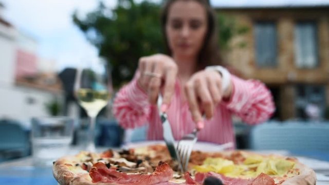 Slow motion of young pretty woman cutting a pizza with knife and fork on a plate. Huge freshly baked Italian pizza with prosciutto, mushrooms and cheese on the table in outdoors restaurant.