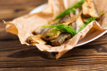 Heap of fried tasty fishes and twigs of fresh green dill on wrinkled paper on white ceramic plate on old wooden rustic brown table