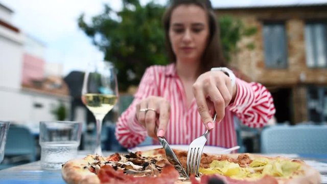 Slow motion of young woman cutting a pizza with knife and fork on a plate. Huge freshly baked Italian pizza with prosciutto, mushrooms, artichokes, tuna and cheese on the table in outdoors restaurant